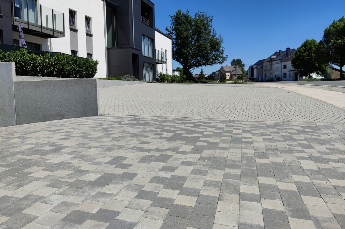 Vue d'une allée pavée moderne dans un quartier résidentiel. Les pavés carrés sont disposés en motifs de différentes teintes de gris, créant un contraste élégant avec l'environnement. La surface de l'allée est divisée en deux zones : une partie en pavés uniformes au premier plan et une partie en pavés avec des interstices pour le drainage à l'arrière-plan.