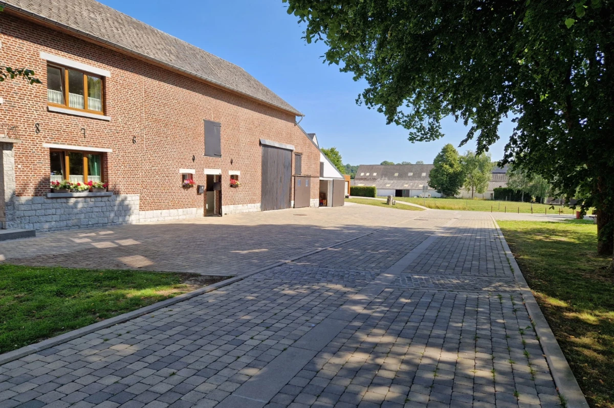 Cour de ferme pavée avec des pavés rustiques en béton tambouriné, mettant en valeur une bâtisse en briques et pierre naturelle dans un cadre rural verdoyant.