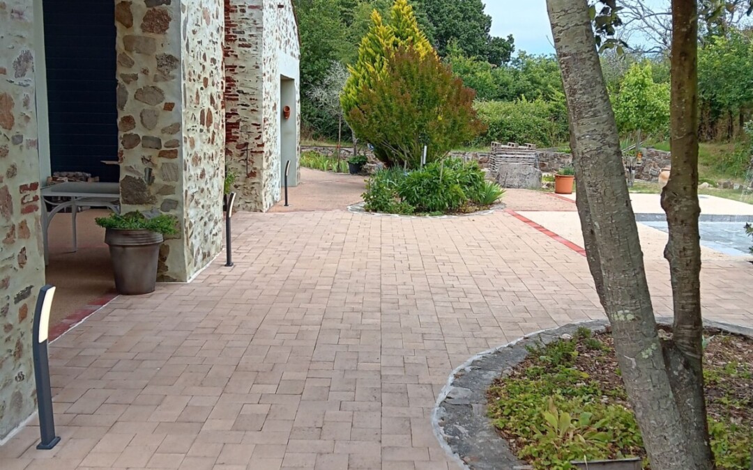 Grande terrasse en pavés francs, coloris clair. La terrasse se trouve le long d'une maison et est bordée de végétation.